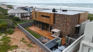 Aerial rear view of House 671 showing timber cladding, stone walls, deck and the ocean beyond